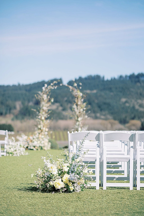 Ceremony setup with white folding chairs facing a floral arch, aisle florals and greenery on a grass lawn under blue sky