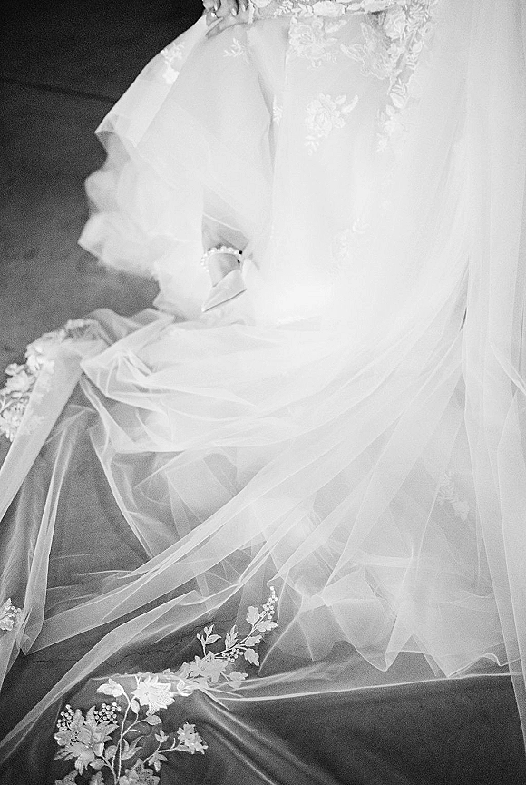 Wedding dress close-up showing tulle wedding dress layers with lace appliqué, embroidered veil, and bridal shoes with rhinestone strap on dark floor