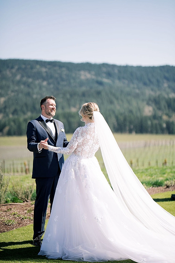 Couple portrait of bride and groom holding hands, her cathedral veil blowing in a sunny meadow with mountain and evergreen forest backdrop