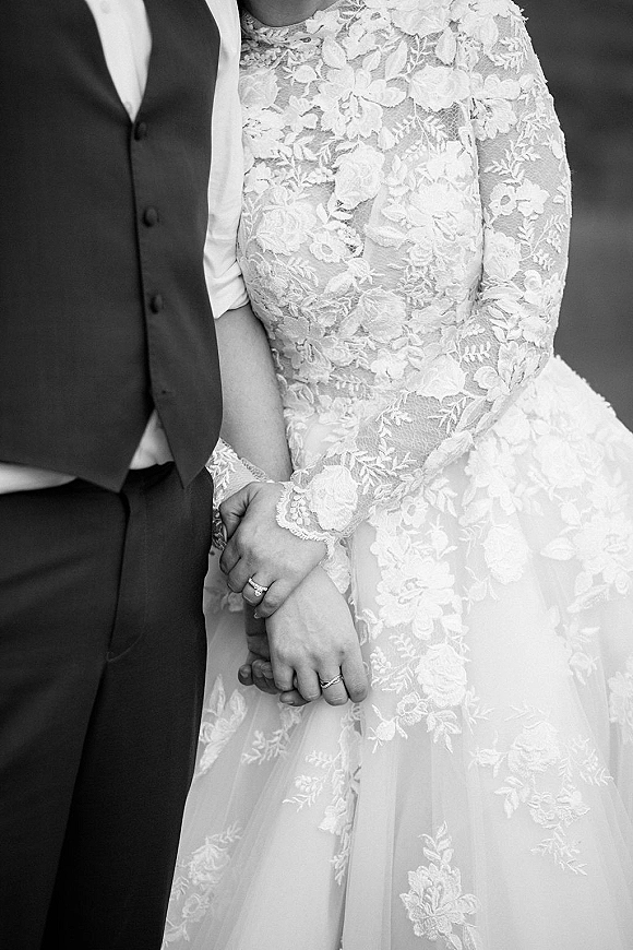 Wedding couple portrait of bride and groom holding hands, highlighting engagement ring and lace long-sleeve gown against a neutral studio backdrop