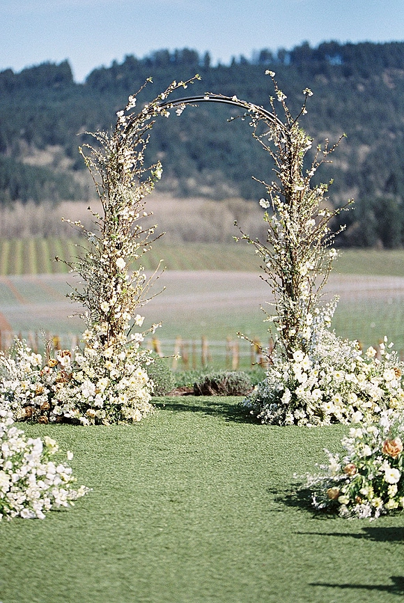 Wedding ceremony arch with white flowers and greenery on a branch frame, set on a grass lawn with meadow blooms and rolling hills behind