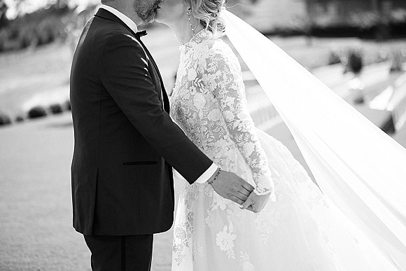 Wedding kiss portrait of the bride and groom kissing as her veil blows in the wind, holding hands on an outdoor lawn with trees and hills