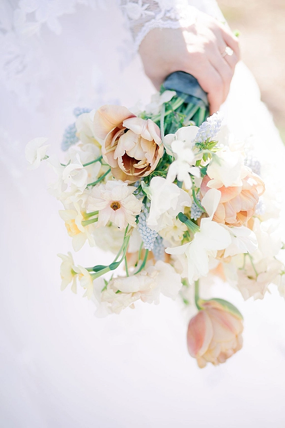 Bridal bouquet in a spring bridal bouquet palette of white and blush blooms with peach roses, held against a white wedding dress with ribbon