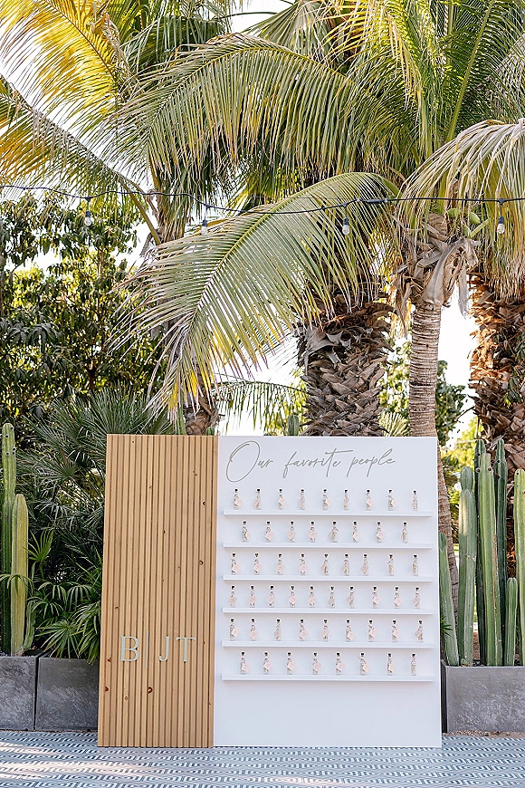 Wedding escort display with an escort card display board featuring mini bottles and calligraphy sign, set on a lit patio with palms and cacti