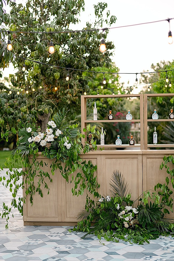 Outdoor wedding bar with wood wedding bar front, back bar shelving of liquor and glassware, trimmed with greenery garland and string lights in a garden setting