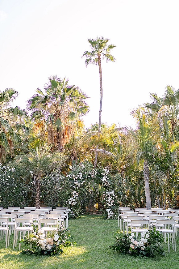 Outdoor ceremony setup with white folding chairs facing a floral arch, aisle florals and greenery on a lawn with palm trees behind