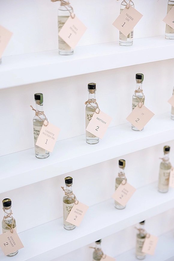 Wedding escort cards with mini liquor bottles and twine-tied name tags arranged in neat rows on white display shelves against a wall