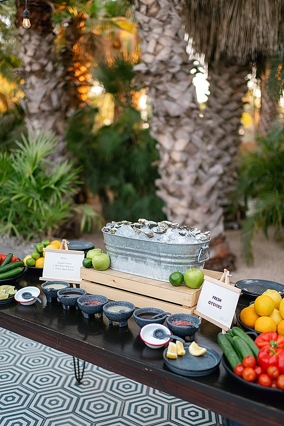 Oyster bar setup with wedding oyster bar signage, oysters on ice in a galvanized tub, citrus and sauces under a hanging bulb in a tropical garden setting