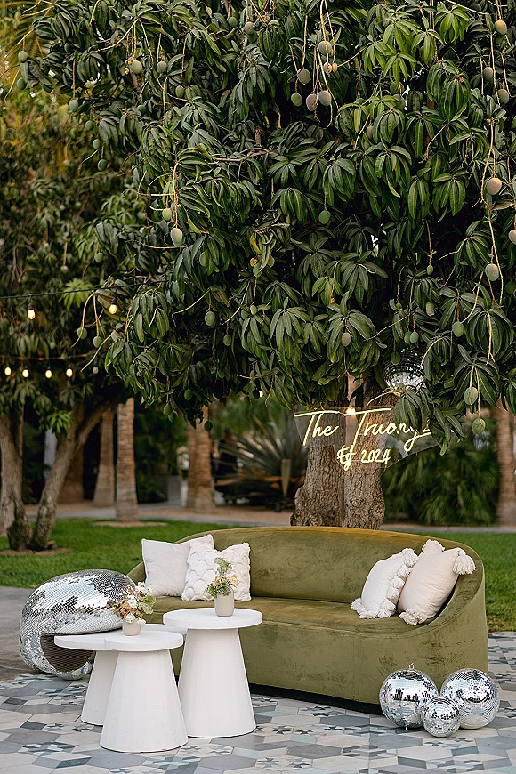 Wedding lounge seating with a green velvet sofa, white pedestal tables and floral arrangements, set under string lights on a garden lawn