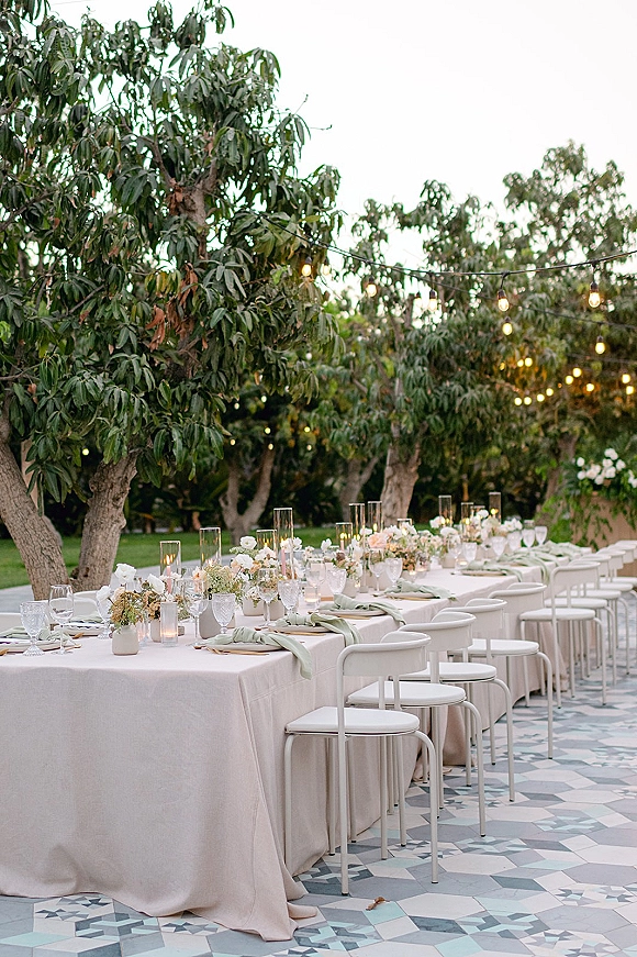 Reception tablescape outdoor reception table with blush tablecloth, sage napkins, floral runners, candles and string lights on a tiled garden patio