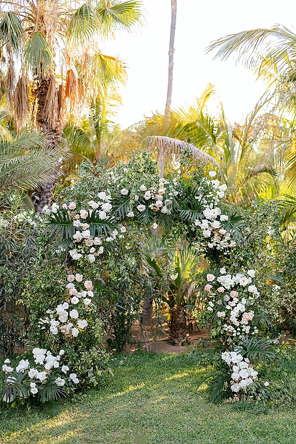 Wedding floral arch circular wedding arch draped with greenery garland, white and blush flowers, and palm fronds in a sunlit tropical garden