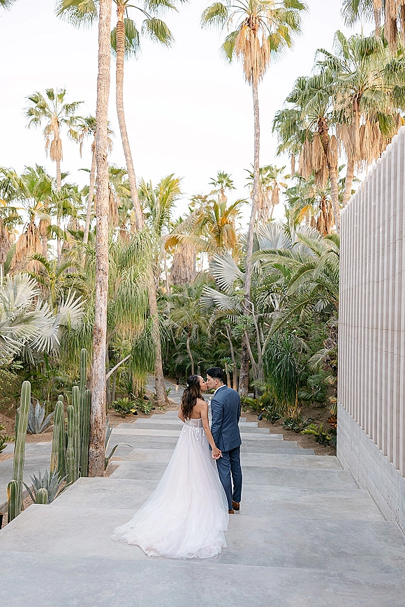 Wedding couple portrait from behind holding hands, bride’s long train and groom in a blue suit walking past palms and cacti on stone steps