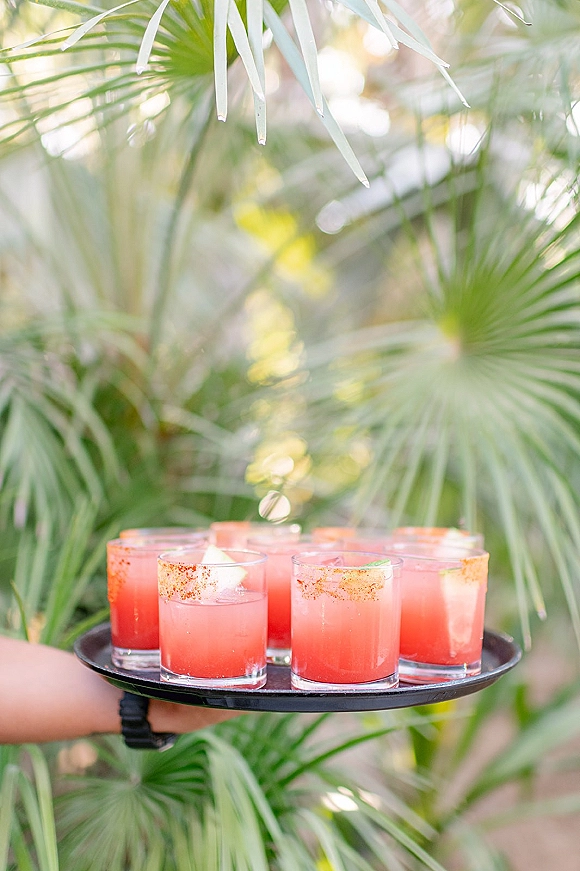 Wedding signature cocktails on a passed cocktail tray, pink drinks with citrus garnish and chili rims, framed by palm leaves outdoors