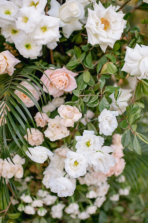 Wedding floral arrangement of white and blush roses with greenery and palm fronds, set against softly blurred foliage backdrop