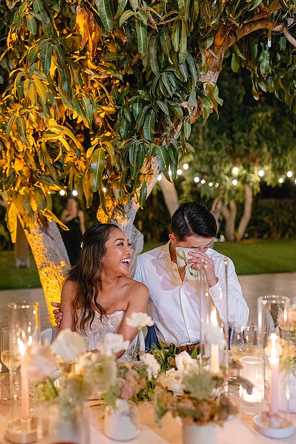 Reception moment at a sweetheart table moment as the bride and groom laugh under string lights, surrounded by candlelit florals outdoors at night