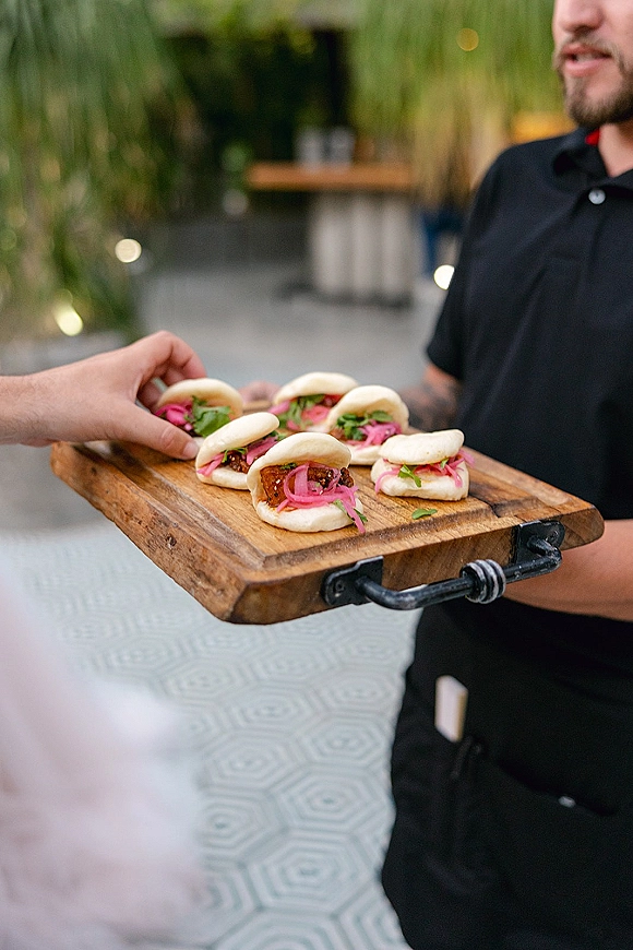 Wedding cocktail hour food with passed appetizers wedding bao buns on a wooden serving board, topped with pickled red onions on an outdoor patio