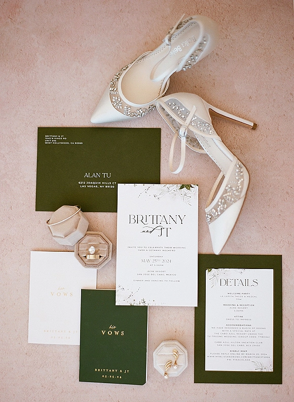 Invitation suite wedding invitation flat lay with RSVP and details cards, dark green envelopes, vow books, rings, pearl earrings on blush fabric backdrop