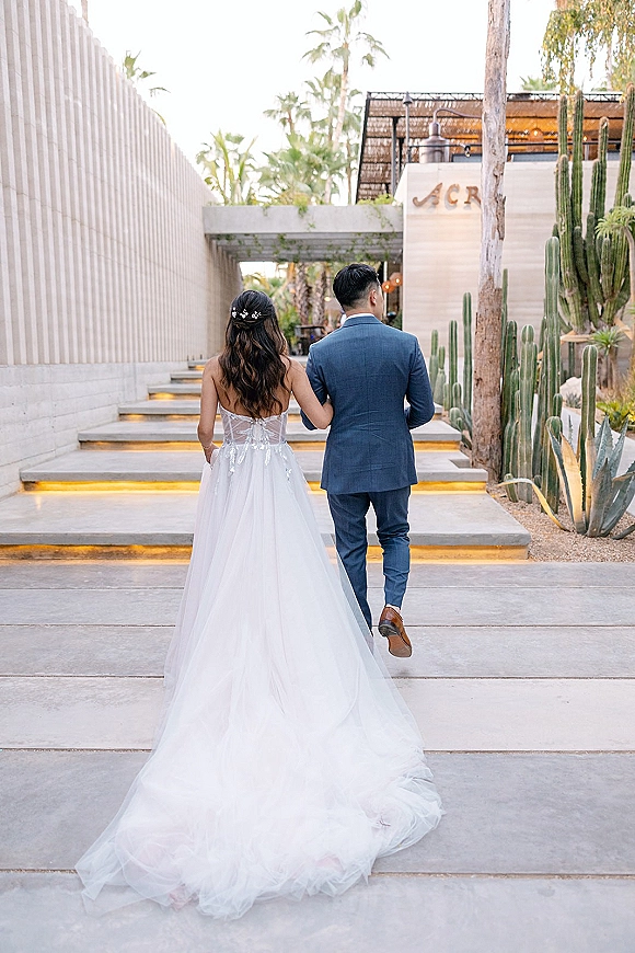 Couple portrait of newlyweds walking away up lit outdoor stairs, bride in a wedding dress with train holding groom’s arm, desert palms and cacti behind