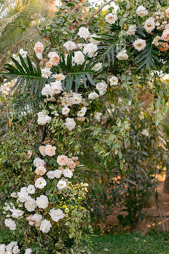 Wedding floral arch with white and blush roses and lush greenery, framed by palm fronds in natural light on a garden lawn