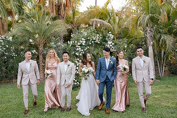 Wedding party photo of bride and groom with bridesmaids and groomsmen walking past palm trees, bouquets and beige suits in a tropical garden