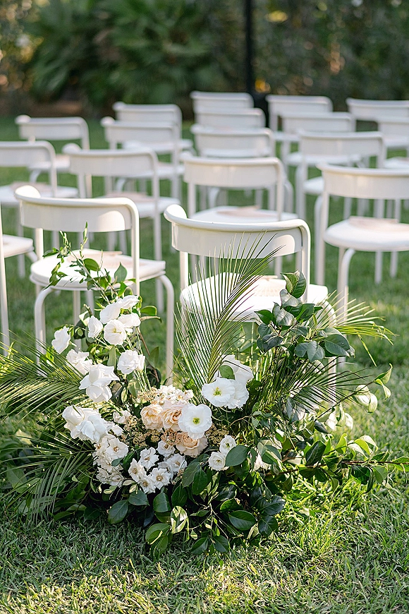 Ceremony aisle decor with white flowers and greenery by white chairs, featuring palm fronds and a low floral arrangement on a garden lawn