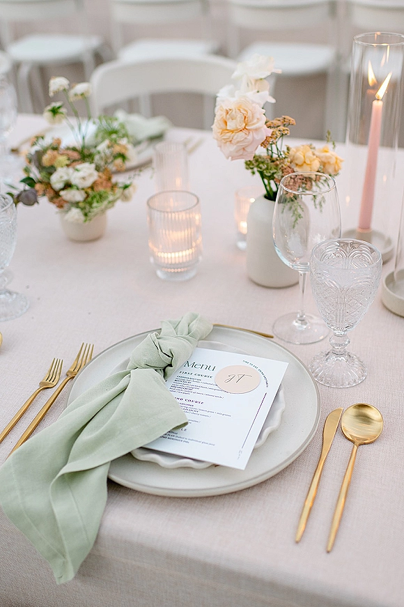 Reception tablescape with wedding place setting, menu card, sage green napkin knot, gold flatware, candles, and pastel bud vases by white chairs