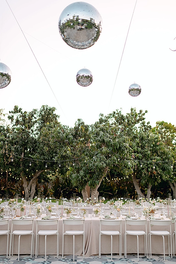 Reception tablescape with outdoor wedding reception details on a long banquet table, low floral centerpieces, string lights, and hanging disco balls in a garden
