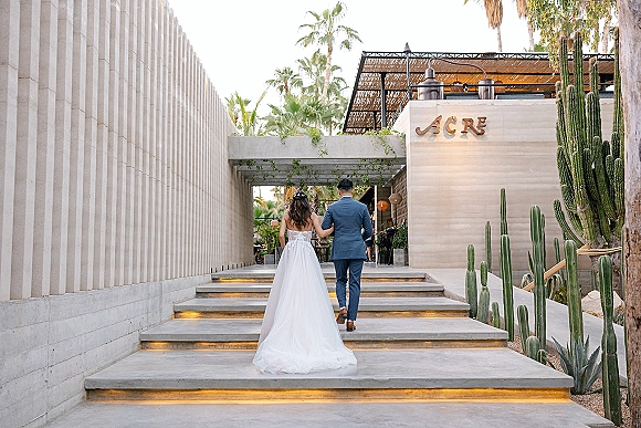 Wedding couple walking up stairs, bride holding her long train in a strapless gown beside groom in a suit at modern venue entrance