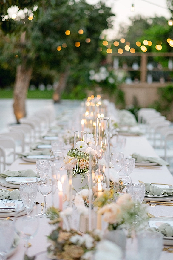 Reception tablescape on a long banquet table wedding with white tablecloth, floral centerpieces, taper candles, and string lights in a garden setting