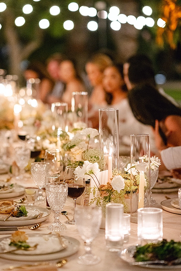 Reception tablescape with an outdoor wedding dinner table set with taper candles in hurricane vases, low florals, and string lights above guests
