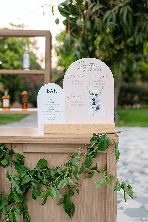 Bar signage with a signature drinks sign on a wooden bar, framed by greenery garland and liquor bottles, set outdoors by trees