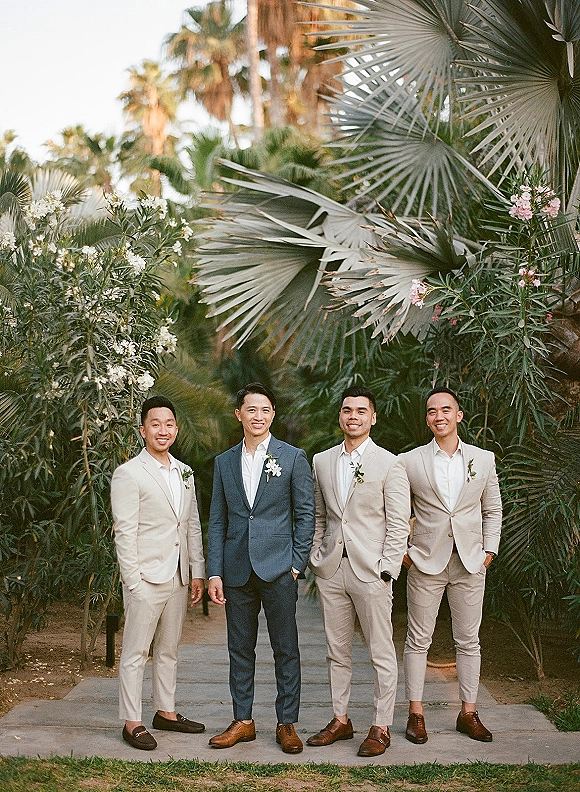 Groomsmen portrait with groom in a blue suit and beige suits, boutonnieres and open collars, walking a palm-lined garden path