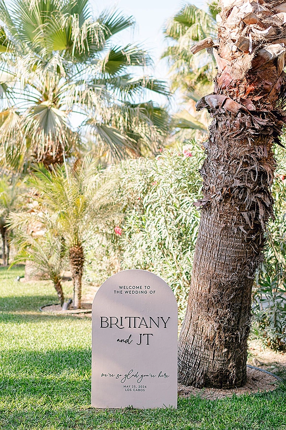 Wedding welcome sign with arched shape and printed typography standing on a sunny lawn amid palm trees and tropical greenery