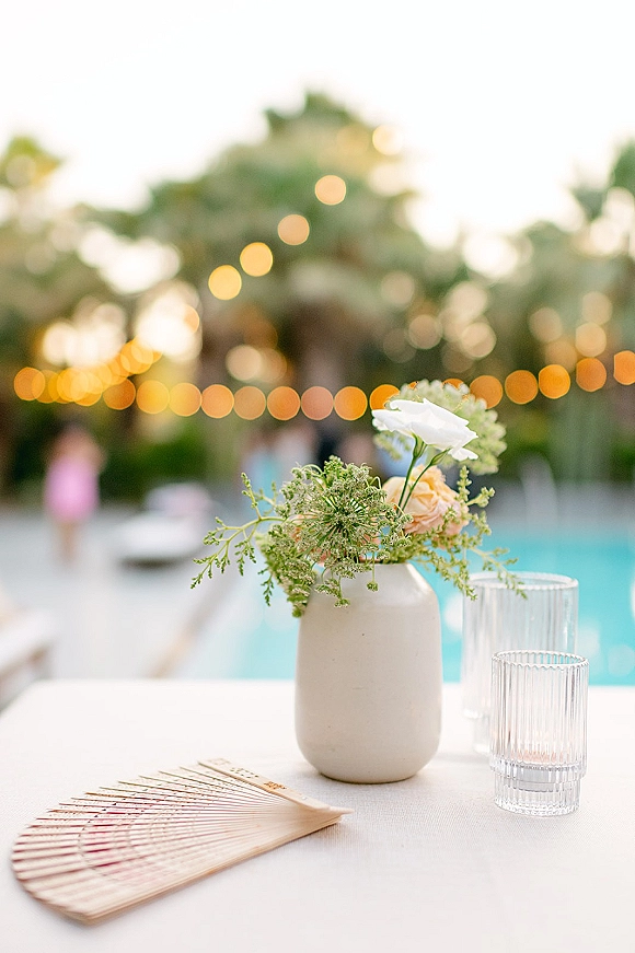 Wedding table centerpiece in a ceramic vase with peach and white flowers, ribbed glassware and a hand fan, poolside under string lights