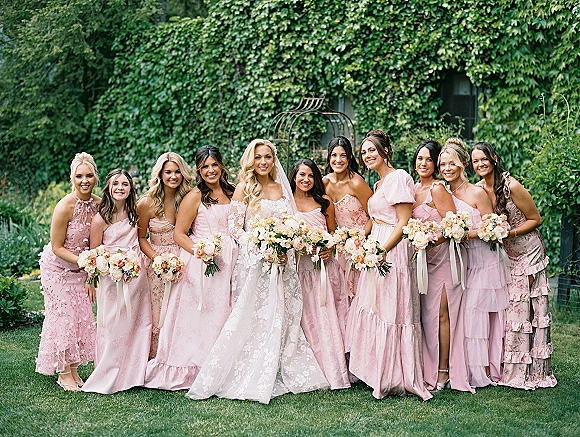 Bridesmaid group photo with bride with bridesmaids in blush dresses holding bouquets, bride in lace veil before an ivy-covered wall