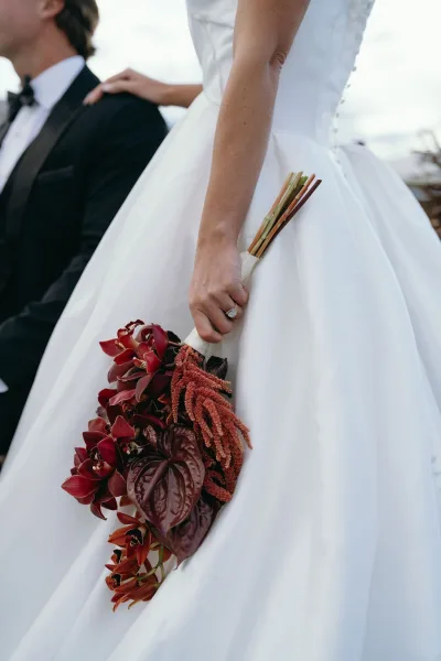 Bridal bouquet in burgundy wedding bouquet tones with cascading orchids and anthurium, held by bride in white gown beside groom in tux under open sky