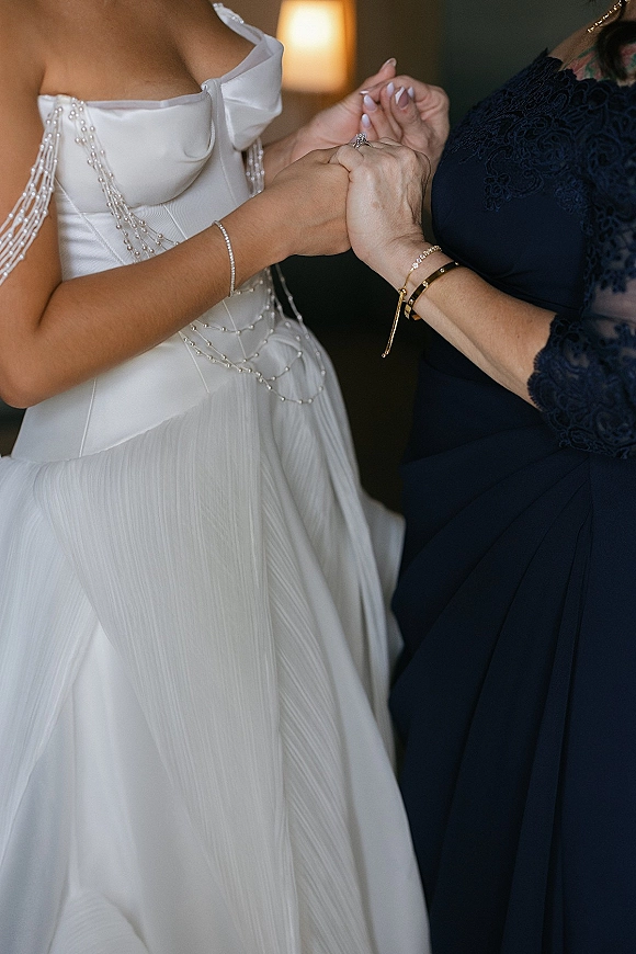 Bride getting ready as her mother holds her hands, highlighting pearl draped straps and wedding ring in warm lamp-lit room