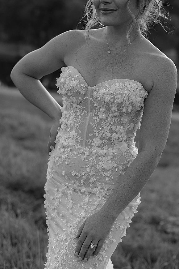 Wedding dress close-up of a strapless lace wedding dress with floral appliqué bodice, pendant necklace and rings, in a blurred field backdrop