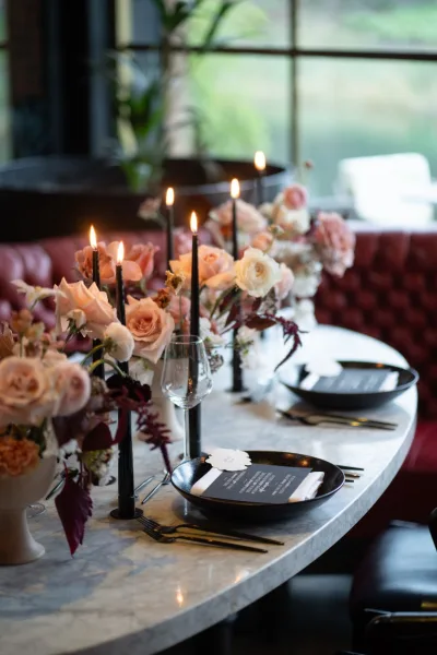 Wedding tablescape with blush rose centerpieces and black taper candles on a marble tabletop, set with black plates, menus, and wine glasses