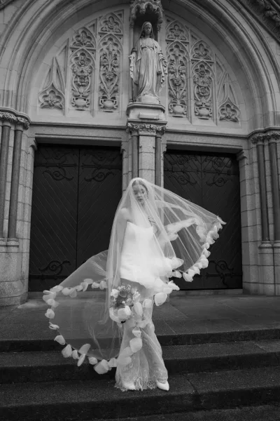 Bridal portrait in black and white of a bride on church steps, veil blowing over her face as she holds a bouquet by stone doors