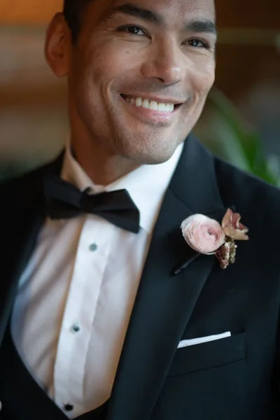 Groom portrait in a black tuxedo with bow tie, white shirt, and pink boutonniere, standing by an indoor wall with greenery
