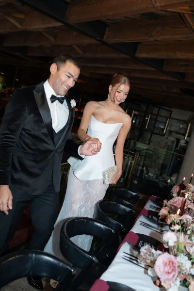 Couple portrait of bride and groom holding hands, entering a restaurant reception beside a long table with burgundy taper candles and florals