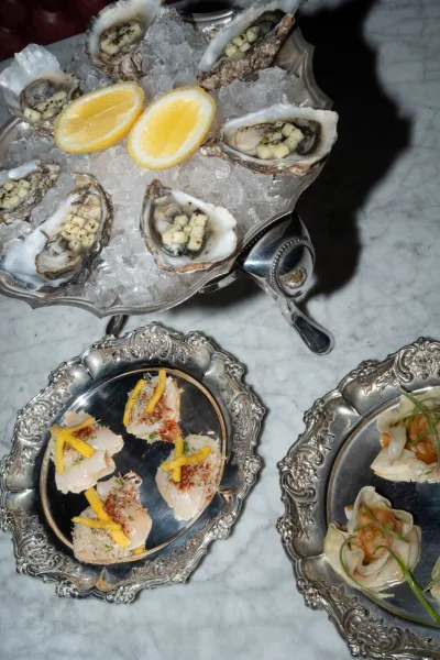 Wedding cocktail hour food featuring an oyster bar wedding spread with oysters on ice, lemon halves, seafood canapés, and dumplings on ornate silver platters on marble table