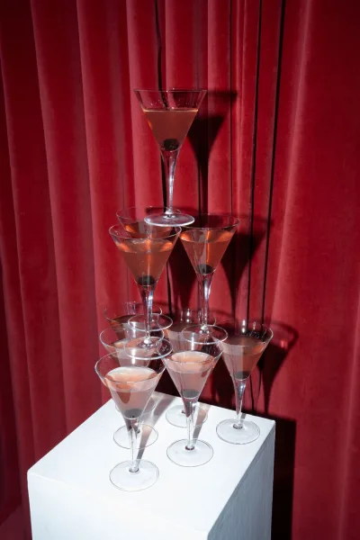 Champagne tower of martini glasses filled with cocktail drinks on a white pedestal, set against rich red velvet curtains backdrop