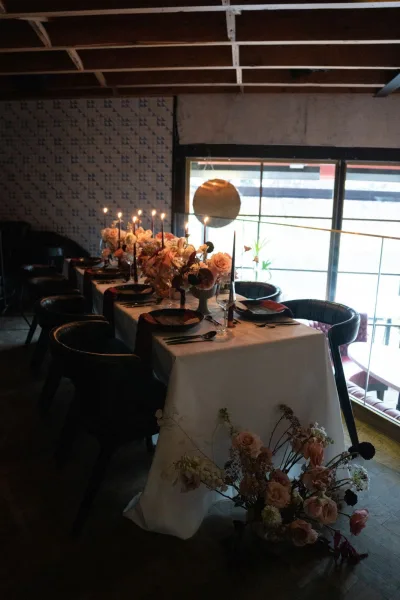 Reception tablescape with wedding head table decor, black plates, floral centerpieces and taper candles on a white tablecloth in a restaurant interior