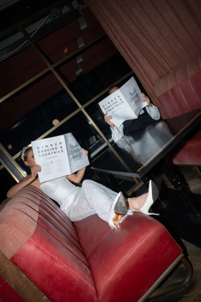 Couple portrait of bride and groom seated reading wedding programs in a leather booth, hiding behind paper in a dark restaurant lounge