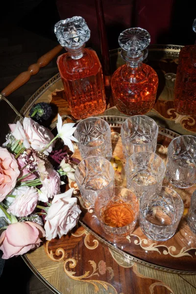 Wedding bar cart with whiskey bar cart crystal decanters and rocks glasses, amber liquor, and a floral bouquet in a dark interior setting