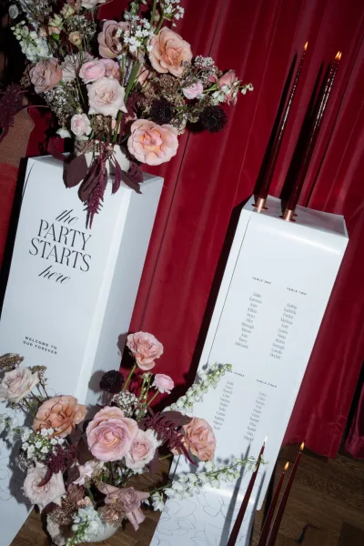 Wedding welcome sign and seating chart on white plinths with blush roses and tall taper candles against a burgundy velvet drape backdrop