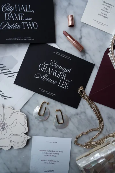 Wedding invitation suite with black wedding invitations and calligraphy lettering, styled with pearls, gold jewelry, lipstick, and wax seal on marble flat lay