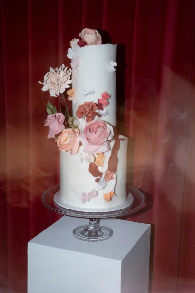 Wedding cake, tiered wedding cake with white buttercream and sugar flowers with a rose topper on a glass stand against red curtains